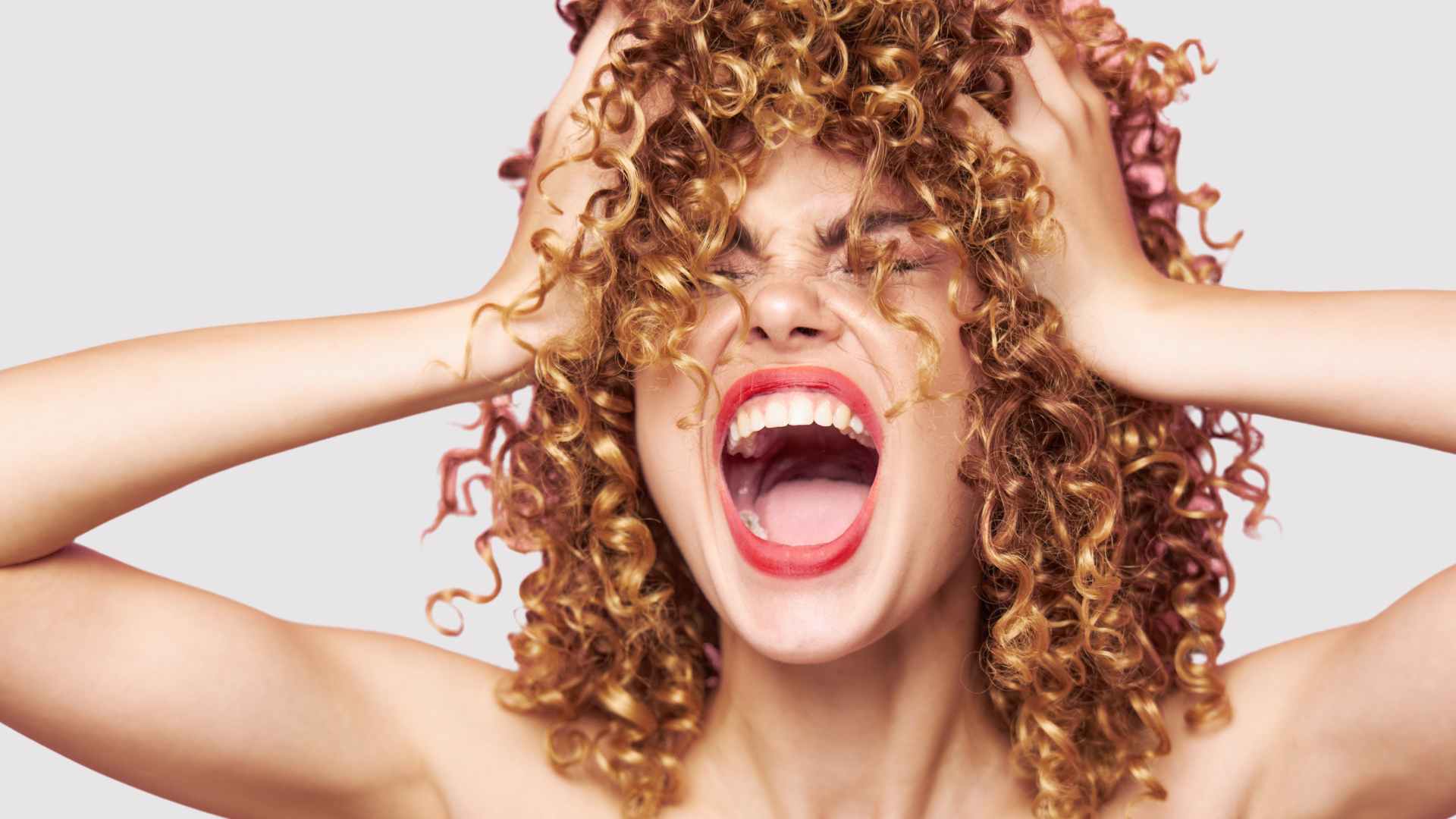 female with shiny hair in curls holding her head in hands and screaming out
